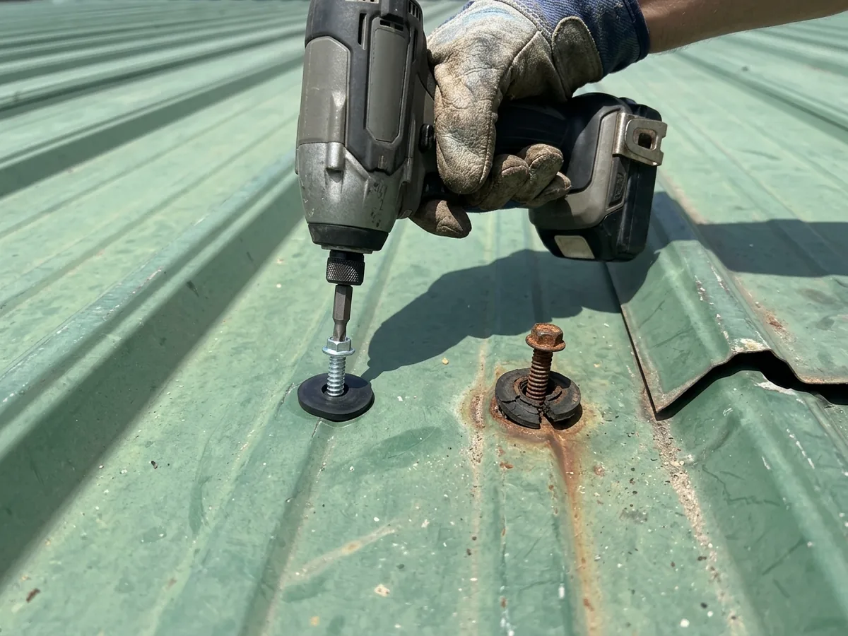 Gloved hand pressing a new screw with a black rubber washer into a ribbed metal roof panel beside a rusted old screw